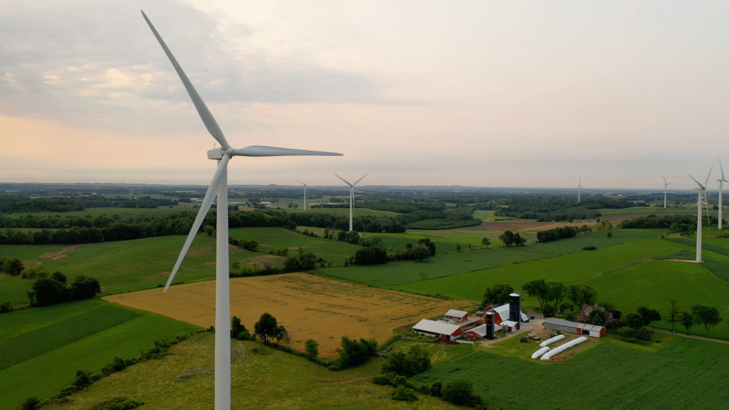 An aerial view of a wind farm in a rural, agricultural setting at dusk. A large wind turbine is prominent in the foreground, overlooking rolling hills with green and golden fields, forests, and a small farm with a red barn.